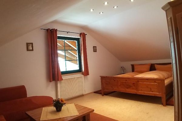 Gemütliches Dachgeschosszimmer mit Doppelbett aus Holz und einer Sitzecke mit Sofa und Tisch. Das Fenster hat rotgemusterte Vorhänge und ein Schrank aus hellem, rustikalem Holz steht seitlich im Raum. Mehrere Strahler leuchten von der Decke.