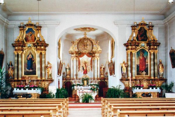 Kircheninnenraum mit zentralem Hauptaltar, davor zwei Seitenaltäre und Bankreihen.