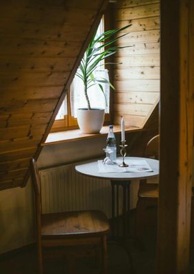 Ein gemütlicher Raum mit holzvertäfelter Dachschräge, mit kleinem Tisch, Stühlen und einer Pflanze am Fenster.