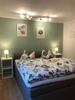 In einem Raum stehen zwei halbhohe, weiße Regale mit Nachttischlampen. Dazwischen steht ein graues Boxspringbett mit grün gemusterter Bettwäsche. An der grün gestrichenen Rückwand hängen zwei Zeichnungen in weißen Rahmen.