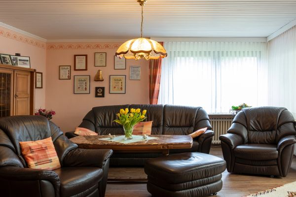 Wohnzimmer mit Ledersofas und Sesseln um einen Tisch, vor einer großen Fensterfront mit Vorhängen und Deckenleuchte.