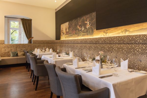 Speiseraum im Restaurant, Tische an der rechten Seite, mit Gläsern, Besteck und Stoffservietten eingedeckt. Warme Erdfarben, Jagdmotive an der Wand.