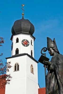 Viereckiger Kirchturm mit eckigem Überbau und Zwiebelkuppel, davor eine Bronzestatue des Heiligen Martin.