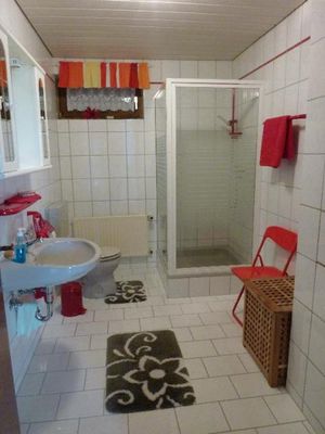 Im hellgefliesten Badezimmer befinden sich eine Viereckdusche mit Glaswänden, WC, Waschbecken, darüber ein Spiegelschrank, ein roter Sessel und ene Wäschetruhe. Der Vorhang ist rot gemustert, Handtücher, Waschbeckenablage, Handtuchhalter und Abfalleimer sind rot.