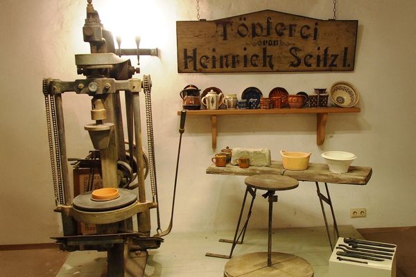 In einem Raum mit weißem Putz stehen alte Töpfereigegenstände. Im Vordergrund ist eine hölzerne Drehscheibe zu sehen, unten eine große und oben eine kleinere, kreisrunde Holzscheibe, die verbunden sind mit einer Metallstange. Daneben steht auf dem Boden ein weißer Würfel, auf dem Metallzangen und andere Hilfsmittel liegen. Dahinter ist eine Holzbank, auf der sich irdene Gefäße und Tonrohmasse befinden. An der Wand dahinter ist ein Holzregal mit verschiedenen bemalten Tongefäßen, worüber ein großes hölzernes Schild mit der Aufschrift "Töpferei von Heinrich Seitz" hängt. Links neben diesem Arrangement steht eine Metallmaschine, die scheinbar für das Formen von Tonblumentöpfen vorgesehen ist.