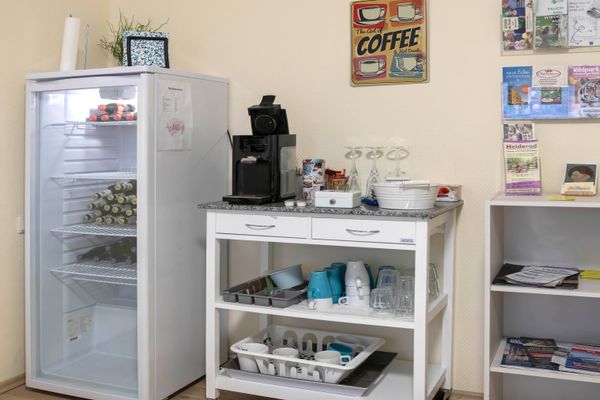Raum mit Getränkekühlschrank, Kaffeemaschine auf weißem Tisch mit Tassen und Gläsern sowie Wandregal mit Prospekten.