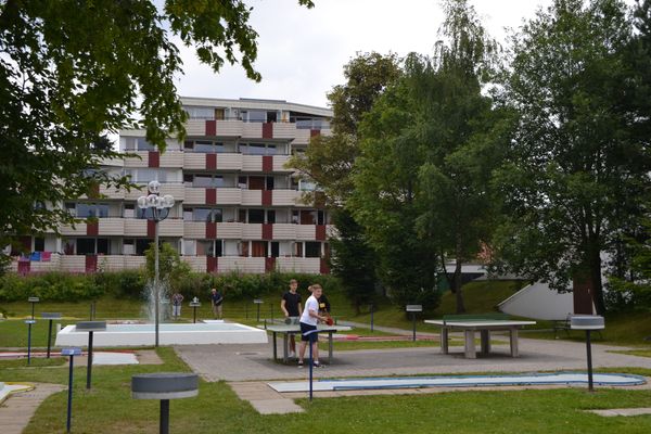 Vor einer Apartmentanlage liegt ein Minigolfplatz mit Springbrunnen und zwei Tischtennisplatten. Zwei Jungen spielen Tischtennis, während eine Frau und ein Mann Minigolf spielen.