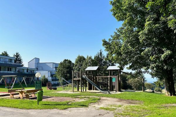 Auf dem Spielplatz vor einer Appartementanlage stehen ein Spielhaus mit Klettergerüst und Rutsche, zwei Schaukelgestelle, sowie ein Sitzbereich mit Holztisch und zwei Bänken. Zusätzlich gibt es eine geschwungene Holzbank. Der Spielplatz wird teils von hohen Laubbäumen beschattet.