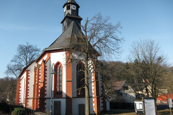 Eine weiße Kirche mit dunkelgrauem Schiferdach und rotem Sandstein an allen Kanten und Fensterlaibungen. Unmittelbar vor der Kiche steht ein kahler Laubbaum. Das Gelände ist mit einer niedrigen Mauer umgeben. Im Vordergrund stehen eine Tafel und Hinweisschilder. Umliegend stehen Häuser. Hinter der Kirche erstreckt sich ein Wald auf einer Anhöhe. Es ist ein klarer schneeloser Wintertag, die vielen Laubbäume sind kahl.