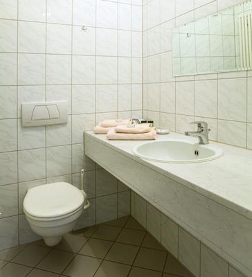 Ein Badezimmer mit hellen Wandfliesen und beigefarbenen Bodenfliesen. Auf einer langen Marmorablage liegen ordentlich gefaltete Bade- und Handtücher. Über dem runden Waschbecken hängt ein breiter Spiegel.