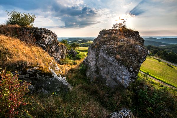 Das Bild zeigt eine hügelige Landschaft mit großen Felsen, die von trockenem Gras und Sträuchern umgeben sind. Auf einem der Felsen steht ein Kreuz, das vom Gegenlicht der Sonne bestrahlt wird, und eine schöne Aussicht auf die grünen Felder im Hintergrund ermöglicht.