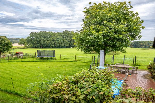 Garten mit Rasenfläche, Terrasse mit Tisch und Stühlen, Baum und eingezäunter Bereich vor offener Landschaft