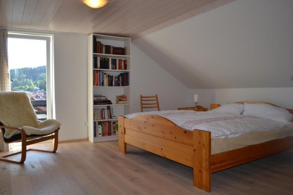 Unter einer Dachschräge steht ein Kieferholz-Doppelbett, daneben ein hohes weißes Bücherregal und ein Stuhl mit Fellauflage; durch die Balkontür blickt man auf den Ort und die bewaldete Hügellandschaft.