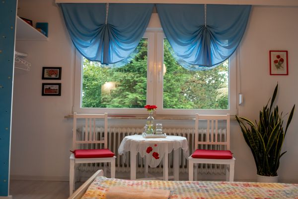 Blick in ein Zimmer auf ein Fenster mit gerafften blauen Vorhängen. Davor stehen zwei weiße Stühle mit roten Sitzkissen und ein kleiner, weiß gedeckter Tisch mit einer Glasflasche und Gläsern. Rechts eine große Pflanze im Topf. Im Vordergrund ist der Anschnitt eines Bettes zu sehen. Durch das Fenster sieht man Bäume.