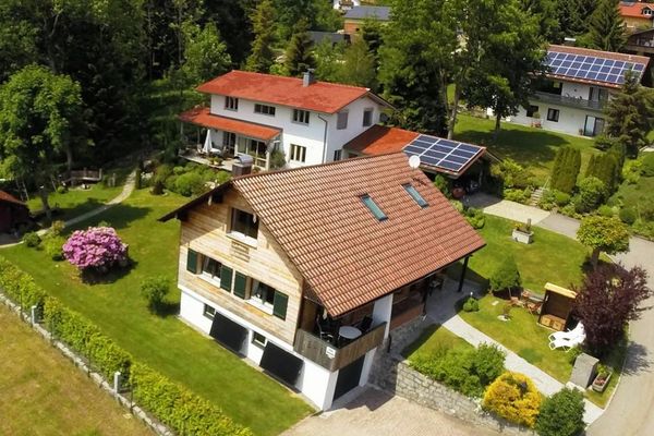 Aus der Luft sieht man das holzvertäfelte Ferienhaus in einem gepflegten Garten mit Pflanzen, Sträuchern, Liegen, Strandkorb und Sitzecke. Dahinter befinden sich weitere Gebäude.