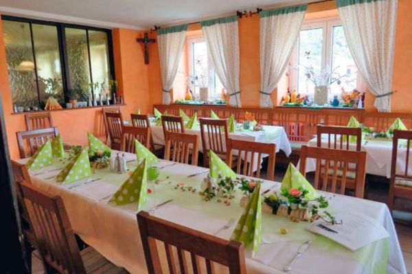 Im Gastraum stehen frühlingshaft gedeckte Tische zwischen Holzstühlen. In der Ecke der orangefarbenen Wand hängt ein Kruzifix. Vor den Fenstern hängen helle Vorhänge mit grünem Muster und Borte. Die Fensterbretter sind österlich dekoriert.