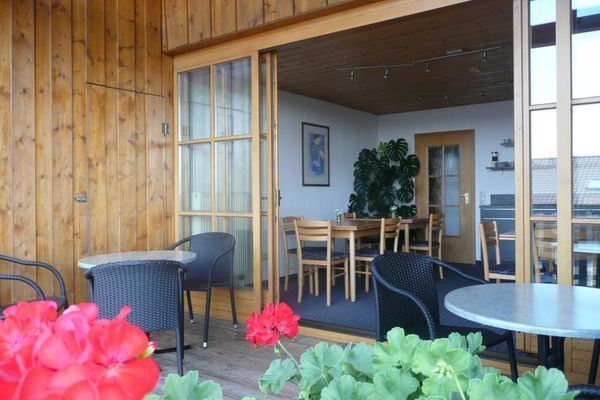 Eine Terrassentürenfront mit Sprossenfenstern verbindet den Frühstücksraum mit der Terrasse. Die Schiebetüren stehen weit offen. Terrassenboden und Hausmauern bestehen aus Holz. Gartenmöbel stehen auf der Terrasse. Im Vordergrund blühen rote Geranien.