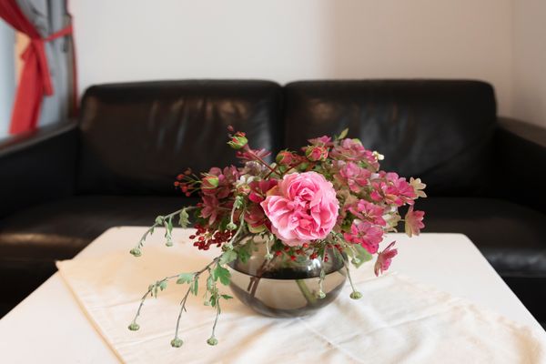 Eine runde, getönte Glasvase mit einem Strauß aus einer großen rosa Rose, kleinen rosa Blüten, grünen Zweigen und roten Beeren steht auf einem weißen Tisch mit heller Stoffdecke
