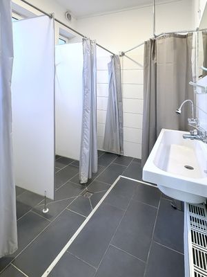 Badebereich mit mehreren Duschen, die durch Trennwände und Vorhänge getrennt sind. Rechts befindet sich ein Waschbecken mit Wasserhahn.