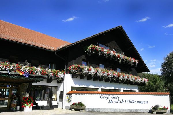 Außenansicht des Hotel-Eingangsbereiches mit üppigem Blumen- und Balkonblumenschmuck. An der gemauerten Gartenmauer steht "Grüß Gott - Herzlich willkommen".