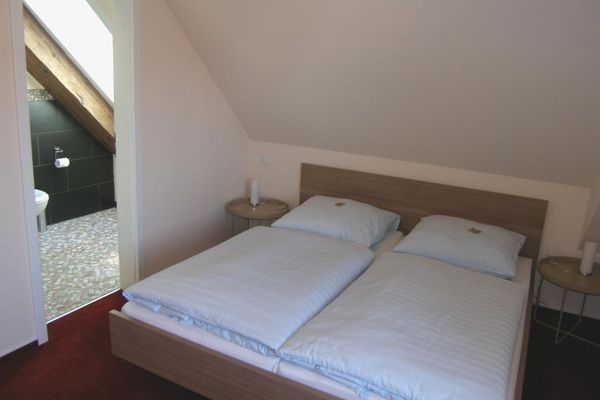 Unter einer Dachschräge steht ein Doppelbett zwischen zwei runden Tischen mit Nachtischlampen. Durch die offenstehende Badtür sieht man den hellen Bodenbelag, die dunkleren Wandfliesen sowie einen Sichtbalken vor dem Dachfenster.