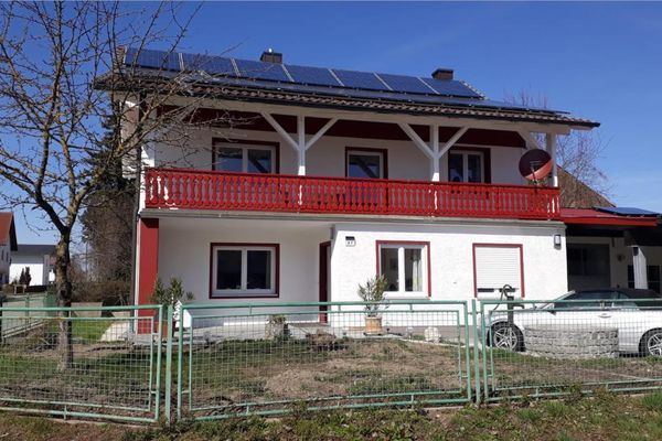Hinter einem grünen Zaun steht ein zweigeschossiges Haus mit einem roten Balkon und einer Photovoltaikanlage auf dem Dach.