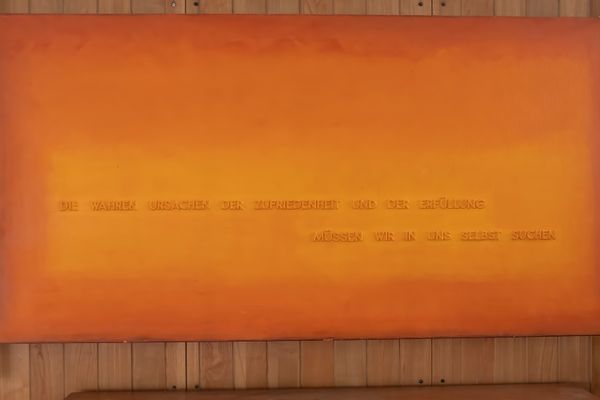 Ein großes, rechteckiges Bild mit einem Farbverlauf in verschiedenen Orangetönen hängt an einer Wand aus hellen Holzpaneelen. Darauf befindet sich mittig ein zweizeiliger Text in erhabenen, orangefarbenen Großbuchstaben.
