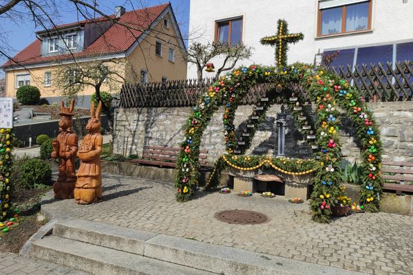 Vor einem Haus erhebt sich die Krone des Brunnens aus grünen Girlanden und bunten Eiern. Seitlich steht eine hölzerne Hasenfamilie.