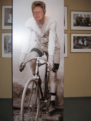 In einem Ausstellungsraum sind alte Schwarz-Weiß-Bilder an der Wand. Ein großes Banner steht davor und zeigt ein altes Bild eines Radreennfahrers. Das Gesicht ist als Oval ausgeschnitten, durch das Loch schaut ein lebendiges Männergesicht.