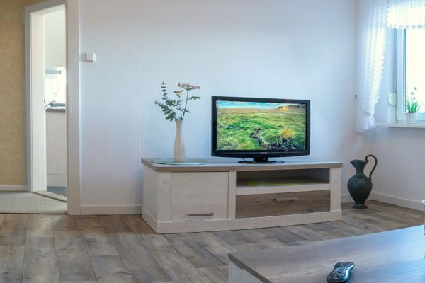 Fernseher auf einem Lowboard, daneben Vase und Fenster mit Vorhang in einem hellen Raum.