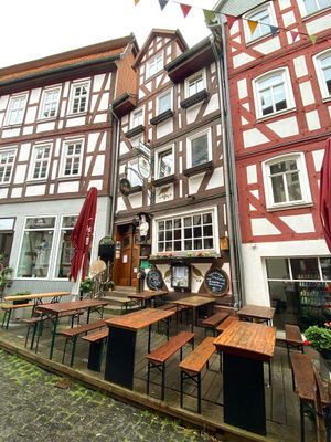 Blick auf ein schmales, hohes Fachwerkhaus, das links und rechts von weiteren Fachwerkhäusern flankiert wird. Durch ein Metallschild über dem Eingang ist schnell zu erkennen, dass es sich um ein Gasthaus handelt. Davor stehen auf einer Holzterrasse einige Brauereigarnituren. Überall sind besondere Details wie alte, runde Backbleche an der Wand zu sehen, die mit weißer Schrift als Speisekarte dienen. Auf dem Gebäude sind in machen Gefachen Wachteln aufgemalt und neben dem Eingang ist die Figur eines dicken Kochs aufgehängt.
