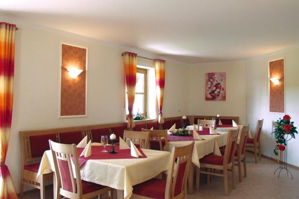 in Aufenthaltsraum mit langer Sitzbank und Stühlen  an mehreren viereckigen Tischen sowie Blumendeko. Sitz-, Rückpolster und Tischmitteldecken sind rot, die Vorhänge rot gemustert. An den Wänden ein Bild mit Blumenmotiv. Zwei Lampen auf orangefarbenem Hintergrund beleuchten den Raum.