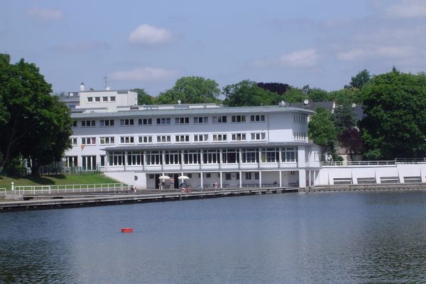 Mehrstöckiges weißes Gebäude am Ufer eines Gewässers mit Bäumen im Hintergrund