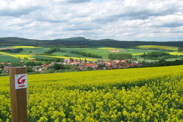 Weiter Blick in eine Frühlingslandschaft über ein üppig gelb blühendes Rapsfeld hinweg. Links steht ein Pfosten mit einer Wegemarkierung, auf der ein rotes G mit Pfeil zu sehen ist. Im Tal hinter dem Rapsfeld liegt ein kleiner Ort, eingebettet in Felder und Wiesen, gesäumt mit Sträuchern und Bäumen. Im Hintergrund sind Anhöhen mit Wald, die den blauen Wolkenhimmel touchieren.