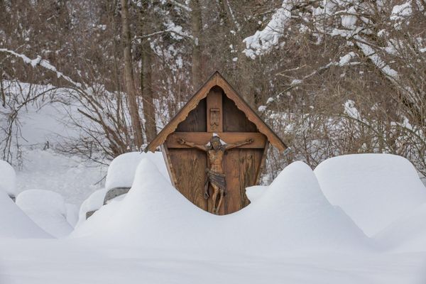 Ein hölzernes, überdachtes Wegkreuz mit Christusfigur ragt halb aus einer Schneewehe; im Hintergrund winterlich kahle Laubbäume.