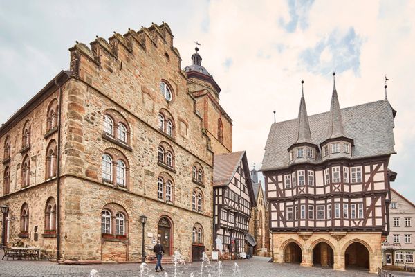 Links ist ein großes steinernes Haus mit fünf Etagen und Stufengibel zu sehen. Daran schließt sich ein kleineres Fachwerkhaus an, hinter dem der steinerne Kirchturm hervorragd. Rechts daneben steht impossant das Fachwerkrathaus auf einer steinernen Bogenhalle gebaut und mit zwei markanten Spitzdach-Gibeln versehen.