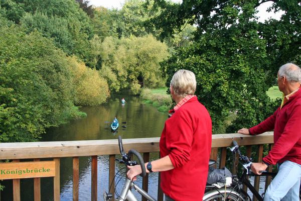 Zwei Radfahrer in roten Jacken lehnen an der Holzbrüstung der Königsbrücke in Bad Bevensen und blicken auf die Ilmenau. Mehrere Paddler fahren flussabwärts durch die von Bäumen gesäumte Landschaft.