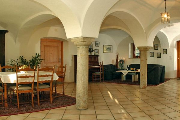 Große Eingangshalle unter einem Gewölbedach, das auf Säulen ruht. Im Sitzbereich ein Zwei- und Dreisitzer-Sofa, Sessel und Tisch sowie ein Klavier. Vor einem wuchtigen Bauernschrank stehen ein Esstisch und sechs Stühle. Der Fliesenboden ist im Sitz- und Essbereich mit Teppichen bedeckt. Mehrere Bilder hängen an der Wand.