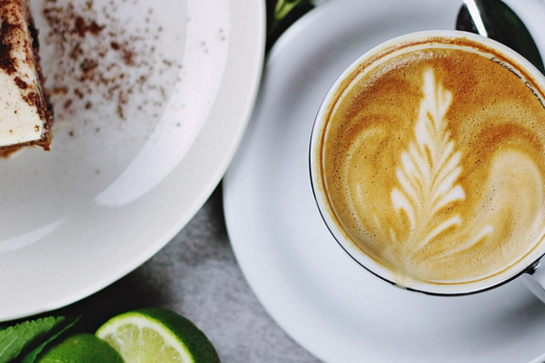 Auf dem Bild ist eine Tasse mit Café darin zu sehen. Daneben liegt ein kleiner Löffel, auf der anderen Seite sieht man einen kleinen Teil eines Tellers mit einem Kuchen.