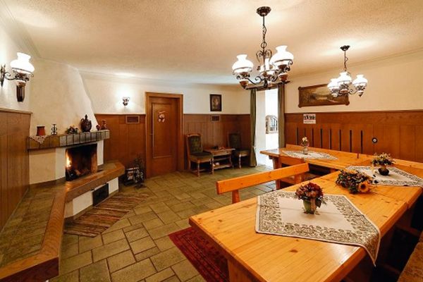 Aufenthaltsraum mit großer Eckbank und Ecktisch, Lampen im Landhausstil, grüner Fliesenboden. Neben der Tür eine Zweier-Sitzgruppe mit Holztischchen. Die Wände sind halbhoch holzverkleidet. Im offenen Kamin brennt ein Feuer.