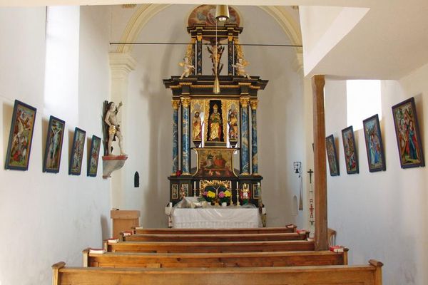 Kleiner Altarraum mit einreihigen Kirchenbänken. An den Seitenwänden Kreuzwegstationen, links eine Heiligenfigur.