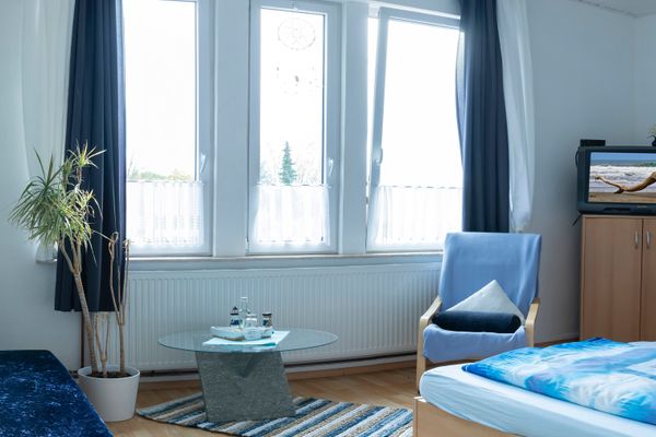Helles Zimmer mit Bett, Sessel, Glastisch und Fernseher vor großen Fenstern