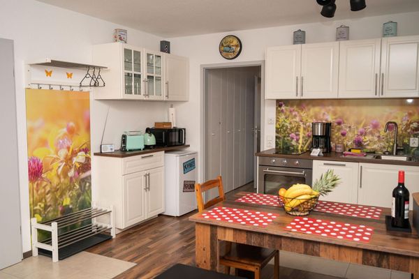 Eine helle Küchenzeile mit Elektrogeräten und einer Blumenmotiv-Rückwand wird durch eine offenstehende Tür geteilt. Ergänzt wird der Raum durch einen Holzesstisch, eine weiße Garderobe mit Schuhablage und Einbauschränke im Flur.
