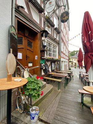 Blick auf den Eingangsbereich eines Fachwerkhauses, das von weiteren Fachwerkhäusern flankiert wird und in einer schmalen Gasse steht. Durch ein Metallschild über dem Eingang ist schnell zu erkennen, dass es sich um ein Gasthaus handelt. Auf einem schwarzen Metallschild an der Wand steht Zur Wachtel mit goldener Schrift geschrieben. Davor stehen auf einer Holzterrasse einige Brauereigarnituren und zugeklappte Sonnenschirme. Überall sind besondere Details wie alte, runde Backbleche an der Wand zu sehen, die mit weißer Schrift als Speisekarte dienen. Auf dem Gebäude sind in machen Gefachen Wachteln aufgemalt und neben dem Eingang ist die Figur eines dicken Kochs aufgehängt. Im Hintergrund, die Kopfsteinplasterstraße hinunter, steht ein Backocker Prunkbau.