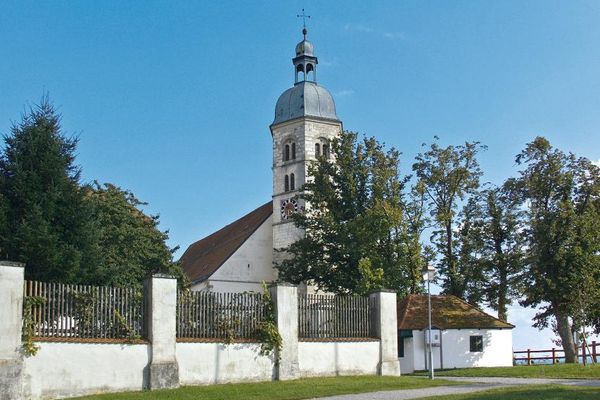 Turm der Wallfahrtskirche Bogenberg, teils von hohen Laubbäumen verdeckt. Davor eine niedere Mauer mit vier hohen, gemauerten Posten und Holzzäunen dazwischen sowie ein kleines Gebäude.