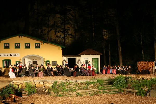 Bei einer Szene im Freilichtspiel sind im Biergarten vor einem großen Wirtshaus alle Tische besetzt von Männern, Frauen und Kindern in historischen Gewändern.