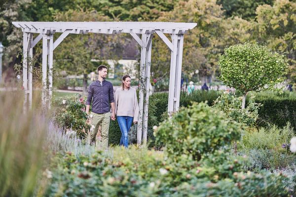 Paar geht unter einem weißen Holzpergola durch einen Garten mit vielfältiger Bepflanzung und Büschen