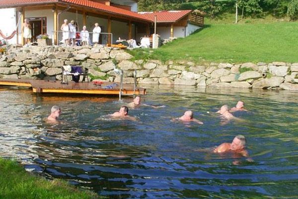Mehrere Damen stehen oberhalb einer Natursteinmauer beim Hotel-Eingangsbereich und sehen den Schwimmern im darunter liegenden Naturbadeweiher zu.