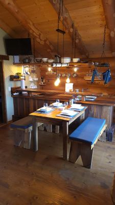 Küche mit Essbereich in einem Holzhaus. Vor dem Küchentresen steht ein Holztisch zwischen zwei Bänken mit Sitzauflagen. An der Wand hängt ein Flachbildfernseher.