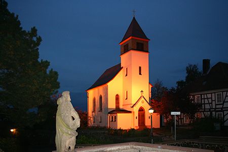 Hell leuchtend strahlt die kleine, weiße Kirche zentral im Bild vor dem dunkelblauen Nachthimmel. Davor ist links eine steinerne Statue zu sehen, die auf einem Sandsteinrahmen eines rechteckigen Brunnenbeckens steht. Am linken Bildrand ist noch eine große Kiefer und rechts steht ein Fachwerkhaus.